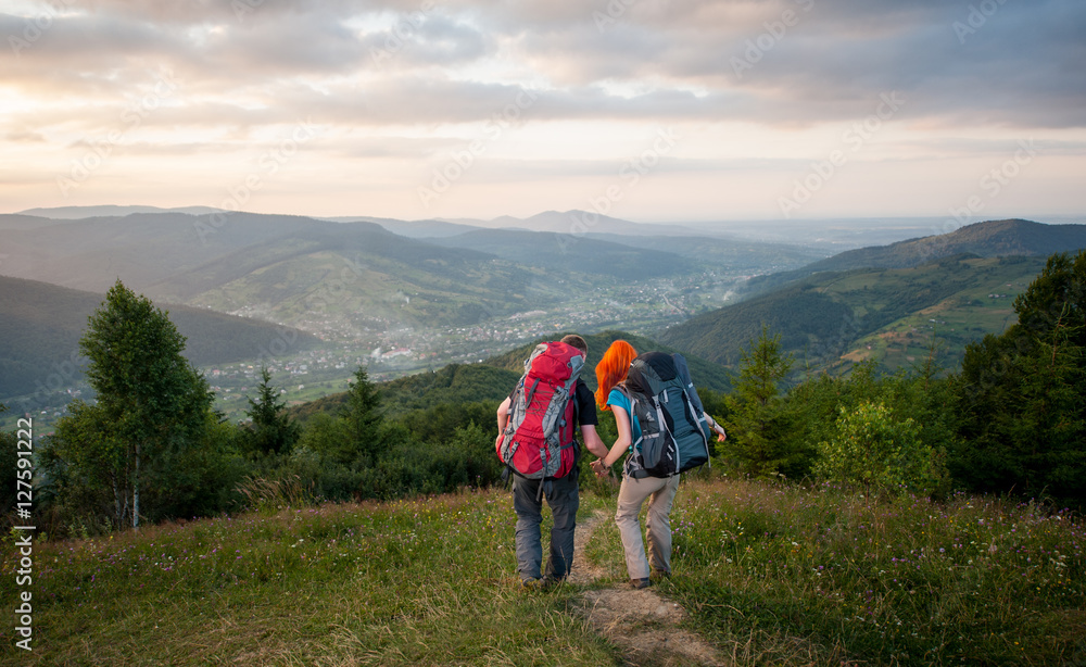 Rear view couple hikers with backpacks walking down from the mountains along the trail. Lifestyle active vacations concept. Mountains, forests, village in the valley, cloudy sky, sunset on background