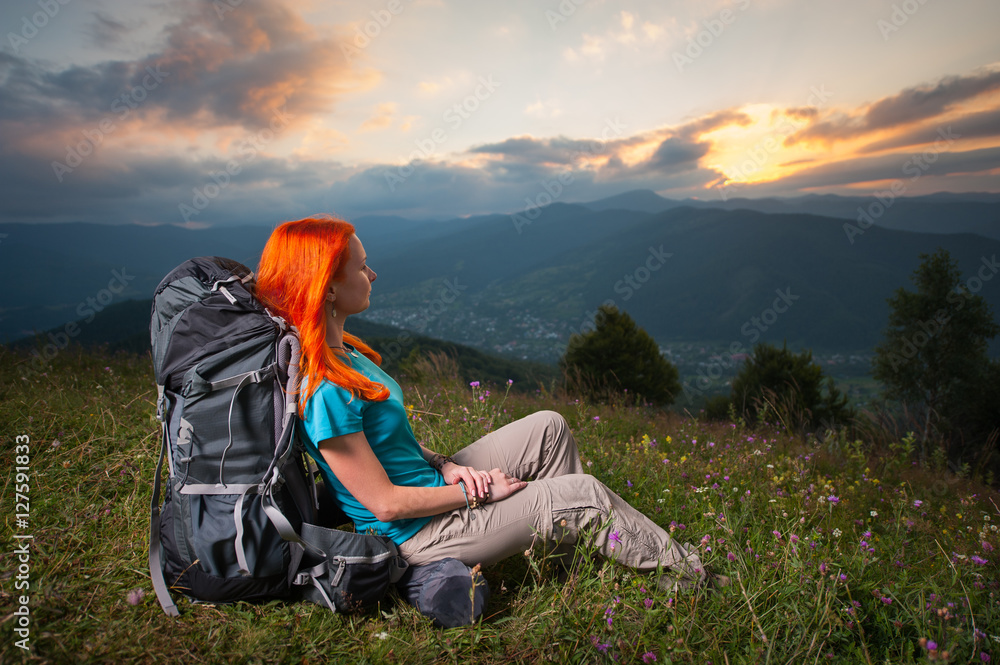 Red-haired girl with backpack sitting in the grass with flowers on a hill and looking over the beautiful mountains, village in the valley and the rays of the sun breaking through the clouds at sunset