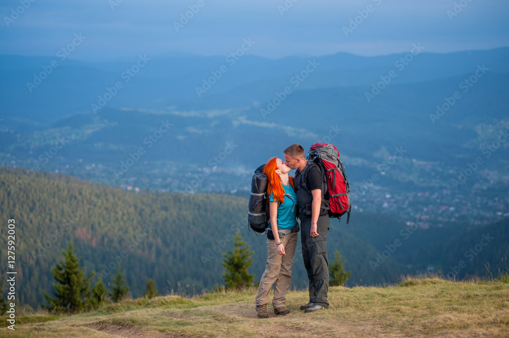 Hikers guy and red-haired woman kissing on the road in the mountain on the background of the landscaped mighty mountains Hikers guy and red-haired woman kissing on the road in the mountain on the background of the landscaped mighty mountains