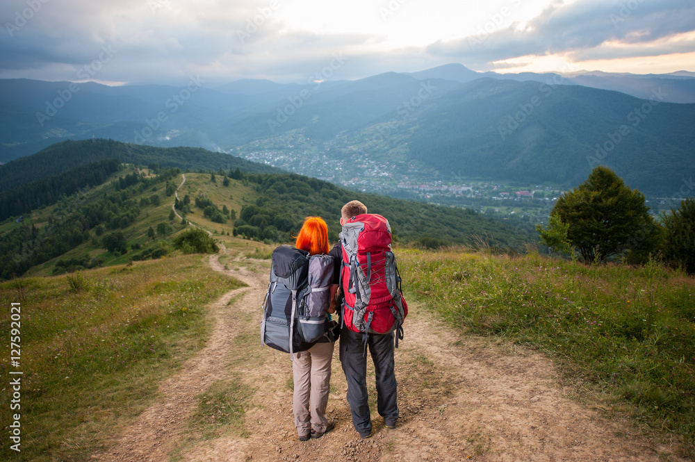 Young couple hikers with backpacks on the road in the mountains. On the background - mountains, forests, hills, village in the valley and cloudy sky