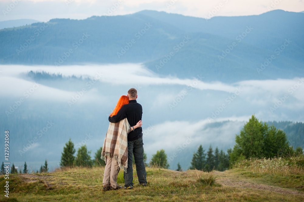 Charming pair standing on a hill enjoying a morning haze over the mountains. Young couple hugging each other. Red-haired girl is covered with a blanket. Rear view