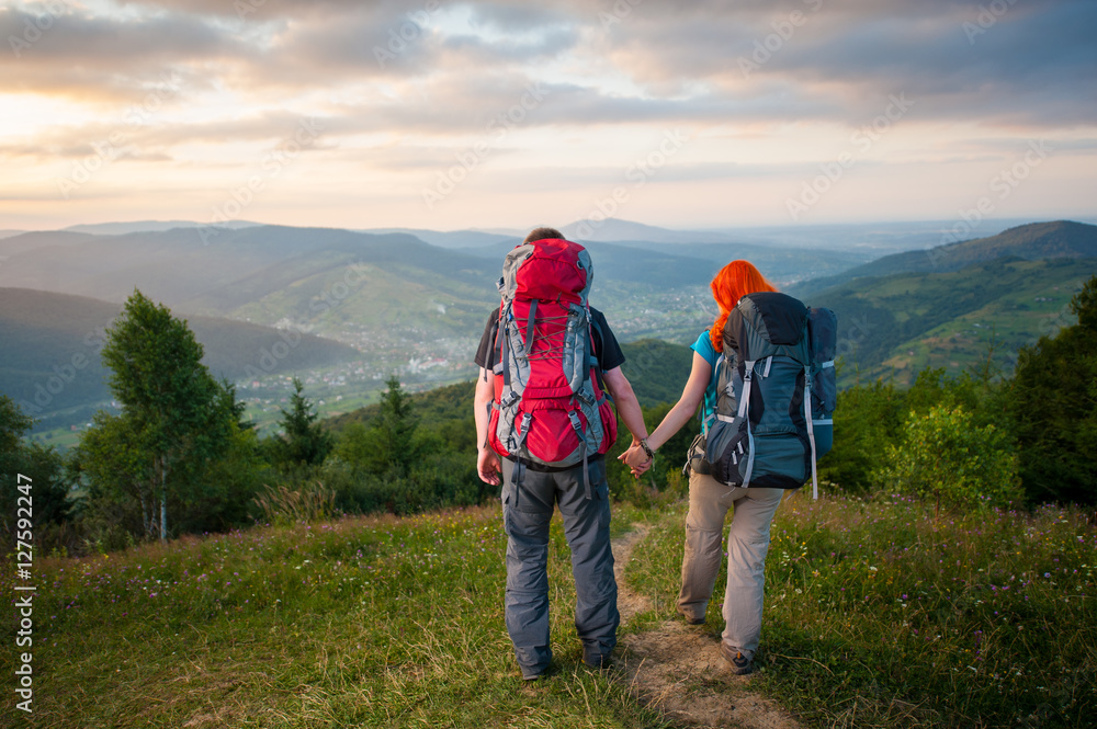 Back view couple tourists with backpacks walking down from the mountains along the trail. Lifestyle active vacations concept. Forests, village in the valley, cloudy sky, sunset on the background