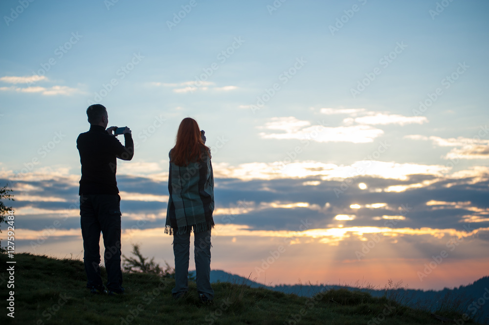 Guy and girl standing on the hill and photographed on phones sunrise. Back view. Beautiful morning in the mountains
