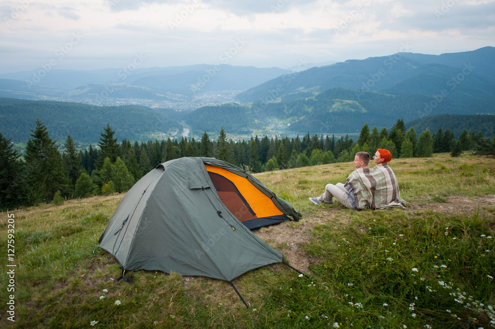 Back view of couple tourists covered with a plaid sitting on a hill near the tent and looking into the distance over the mountains, forest and cloudy sky