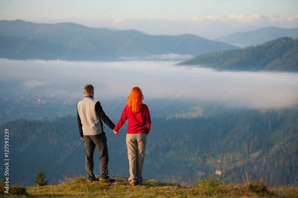 Back view romantic couple standing on a hill, holding hands, enjoying a morning haze over the mountains
