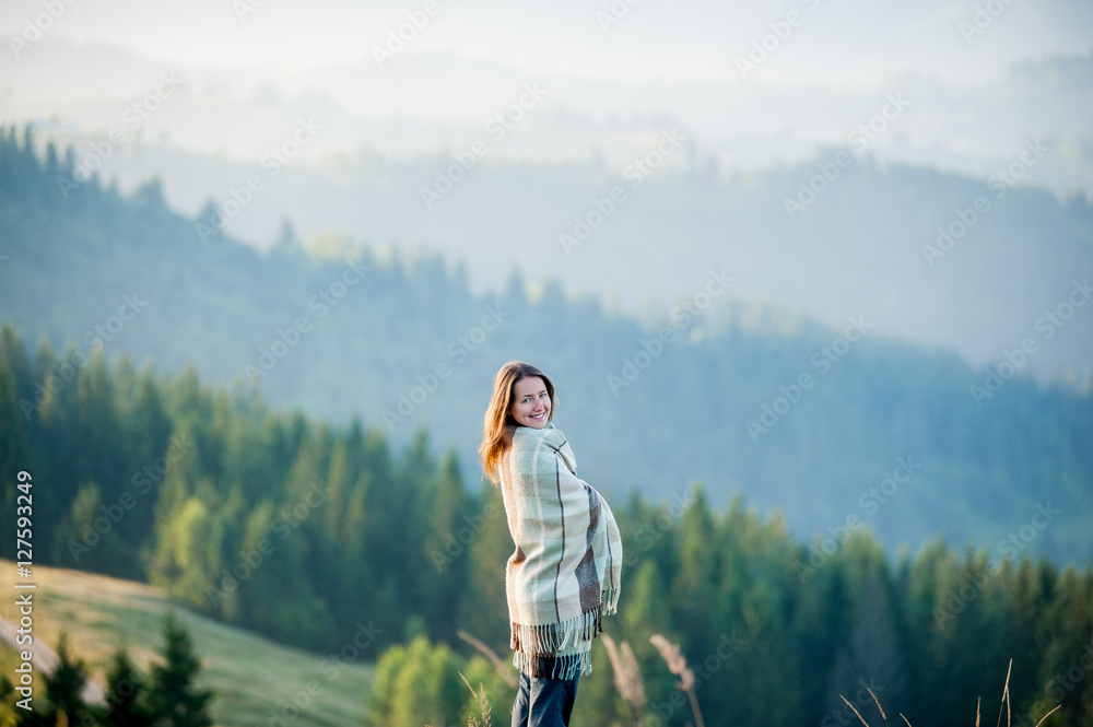 Close-up portrait of happy woman covered with a plaid standing on a hill against beautiful mountain landscape with morning haze over the mountains and forests