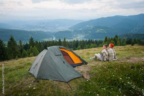 Back view of couple tourists covered with a plaid sitting on a hill near the tent and looking into the distance over the mountains  forest and cloudy sky