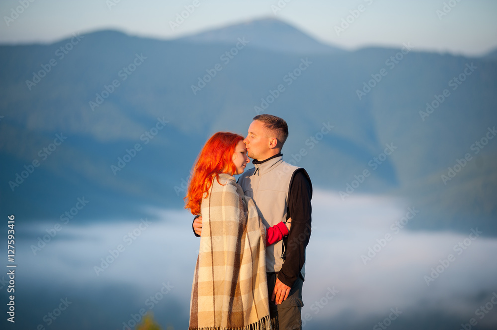 Naklejka premium Romantic couple tourists kissing and hugging each other against beautiful mountain landscape with morning haze over the mountains on background. Red-haired girl covered with a blanket. Close-up