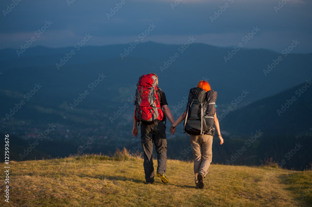 Back view of couple hikers with backpacks holding hands and walk along a road with beautiful mountain landscape on background
