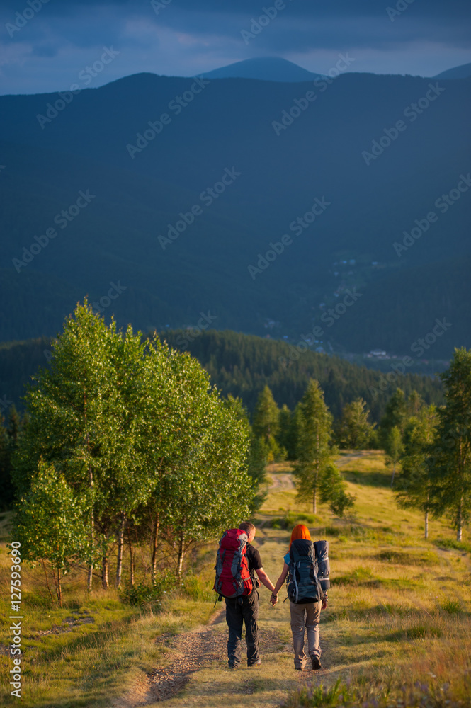 Rear view couple tourists with backpacks walking along a beautiful mountain area holding hands. Lifestyle active vacations concept mountains landscape on background