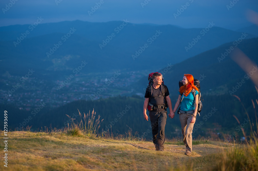 Travelers man and woman with backpacks climbing up in the beautiful mountains area, holding hands and looking to each other. Lifestyle active vacations concept mountains landscape on background
