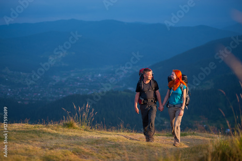 Travelers man and woman with backpacks climbing up in the beautiful mountains area, holding hands and looking to each other. Lifestyle active vacations concept mountains landscape on background