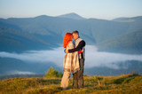Romantic couple standing on a hill, hugging each other against beautiful mountain landscape with morning haze over the mountains on background. Red-haired woman covered with a blanket