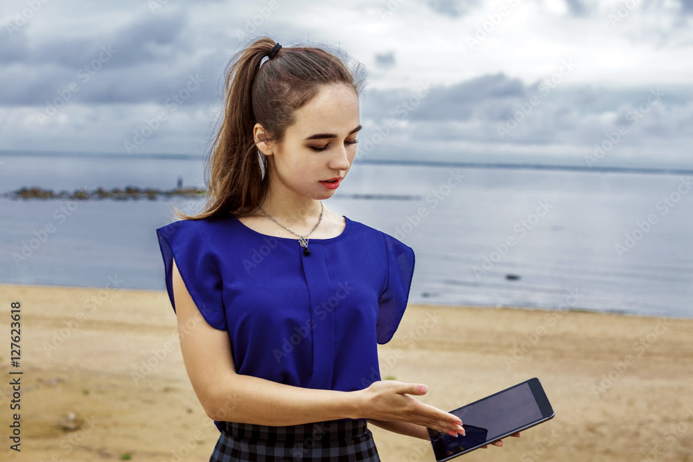Beautiful girl with long brown hair holding a tablet computer. She is dressed in a blue blouse and plaid skirt. Against the background of the beach.