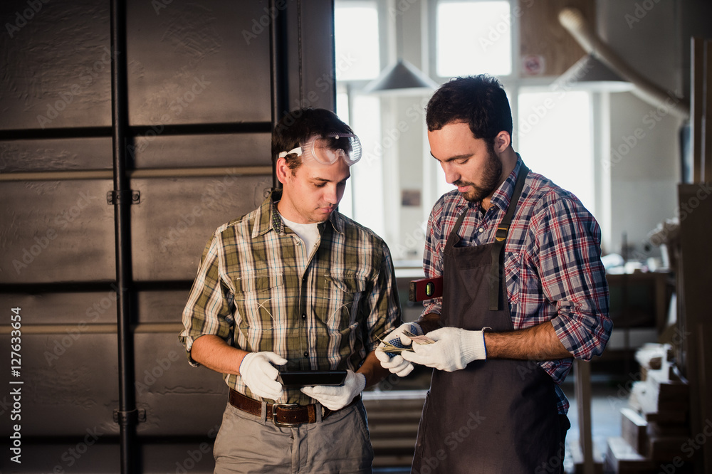 Two carpenters are talking while holding papers and ruler. One of them is wearing safety mask. Carpentry shop background Two carpenters are talking while holding papers and ruler. One of them is wearing safety mask. Carpentry shop background