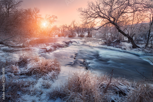 Christmas background with snowy forest. Winter landscape with snowy trees, beautiful frozen river and bushes at sunset. Winter forest. Colorful sky. Water in motion. Trees covered with snow and ice