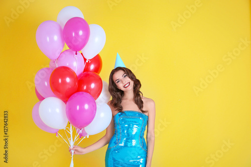 Beautiful young woman in party hat with air balloons on yellow background