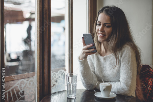 Beautiful woman sitting alone in cafeteria, drinking coffee and looking at cell phone.