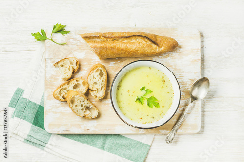 Green vegetable soup with parsley and baguette on white shabby wooden board over white painted wooden background, top view, horizontal composition