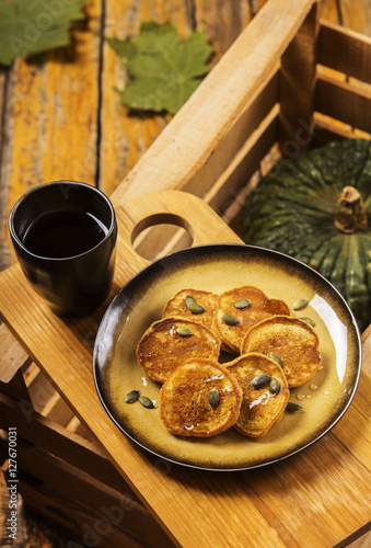Pumpkin pancakes topped with honey and sprinkled with pumpkin seeds on a plate with the effect of craquelure on a wooden box with a cup of coffee and a bowl with honey. Selective focus 