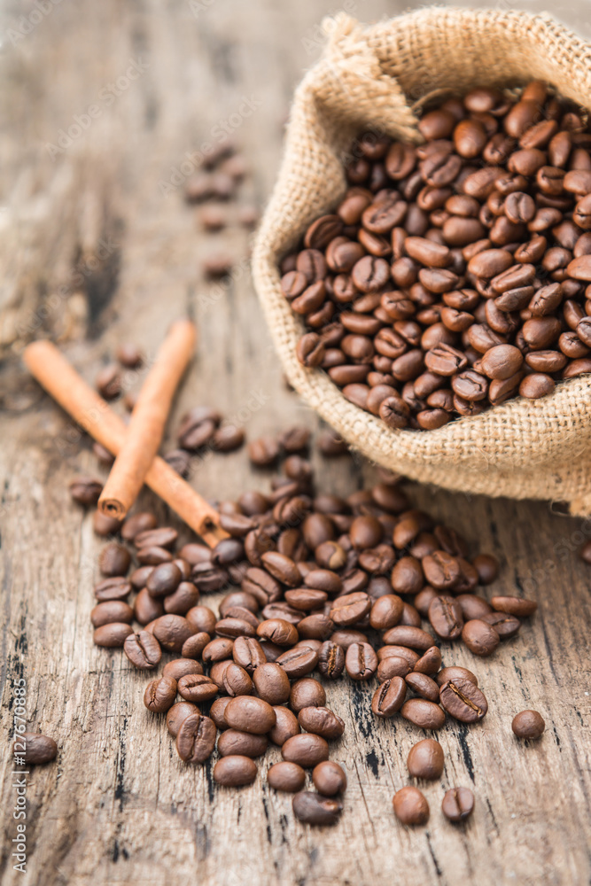 Roasted coffee beans spilling out of the burlap sack bags with cinnamon sticks. Wooden table background. Roasted coffee beans spilling out of the burlap sack bags with cinnamon sticks. Wooden table background.
