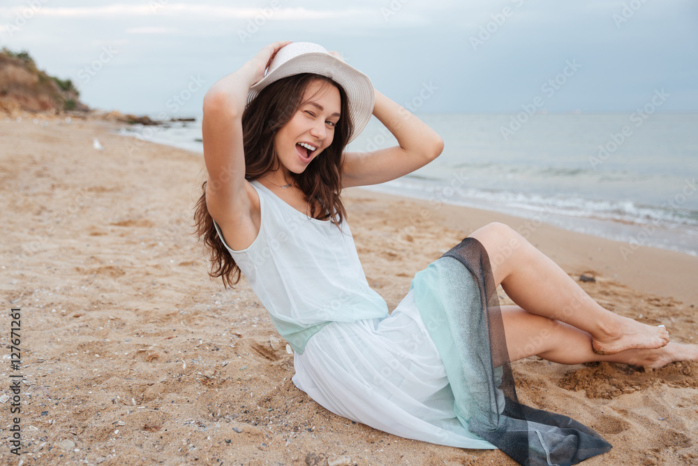 Cheerful playful young woman sitting and winking on the beach Cheerful playful young woman sitting and winking on the beach
