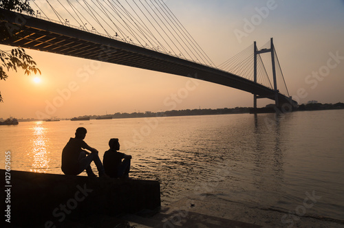 Canvas Print Two friends enjoy the sunset view on river Hooghly with Vidyasagar setu (bridge) at the backdrop at Princep Ghat, Kolkata, India