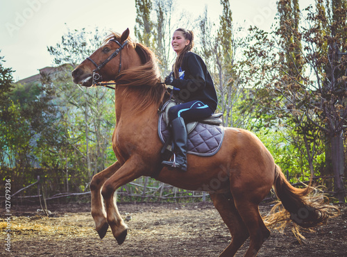 Young beautiful brunette girl rides a horse on a warm and sunny autumn day. Portrait of a pretty young woman on the horse, wearing tall boots and gloves.