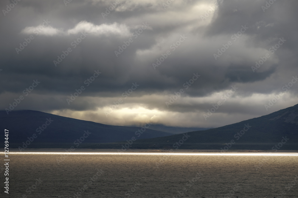 Fototapeta premium Golden colored sunlight at golden hour on large lake with dramatic and moody landscape background