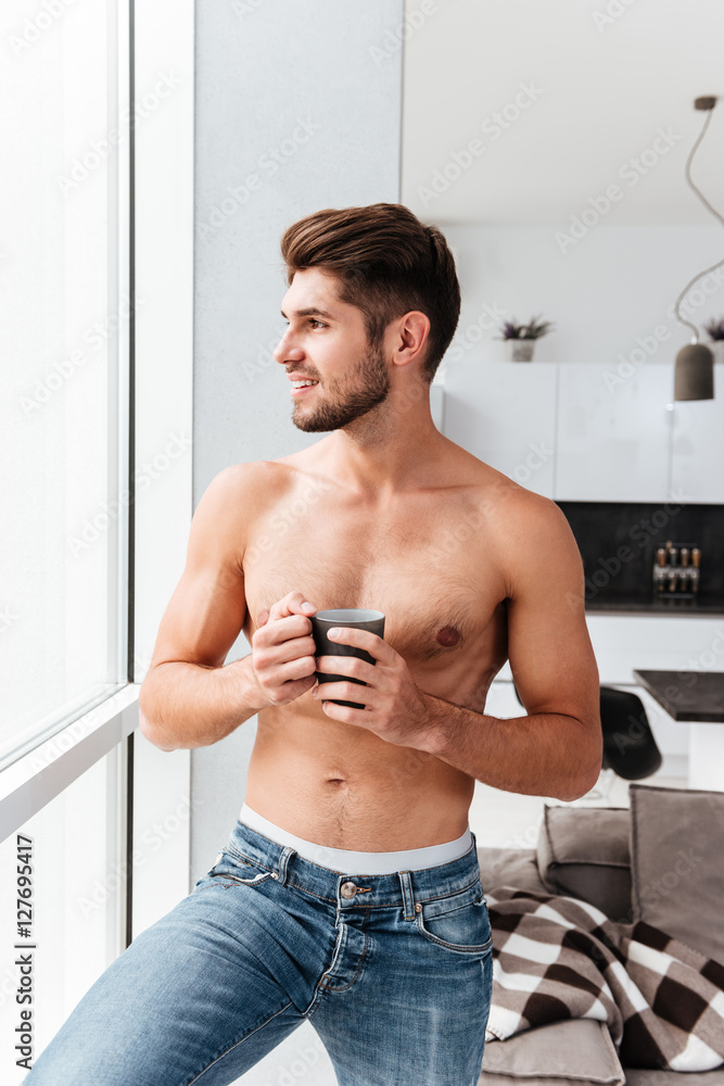 Cheerful shirtless young man standing near window and drinking coffee Cheerful shirtless young man standing near window and drinking coffee