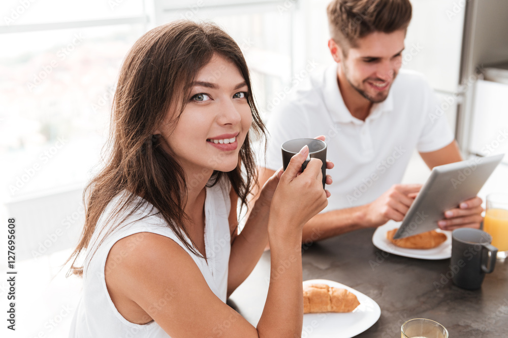 Woman drinking coffee while her boyfriend using tablet on kitchen Woman drinking coffee while her boyfriend using tablet on kitchen