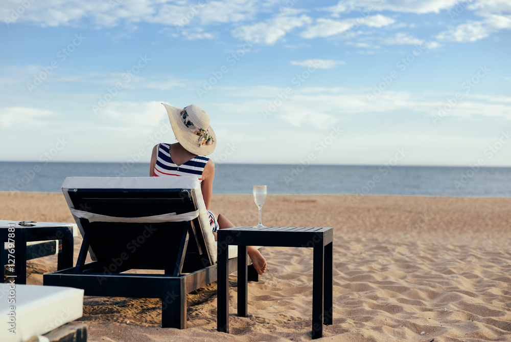 Back view of elegant lady relaxing on a chaise lounge, sandy beach outdoors background