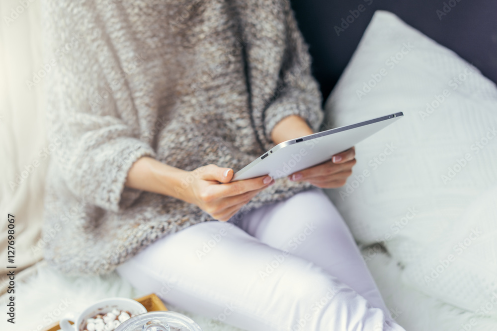 Fototapeta premium Close-up of tablet computer in female hands. Girl, dressed in knitted sweater and white trousers, sitting on white carpet. Near white pillows. Woman using digital gadget. Girl checking email.