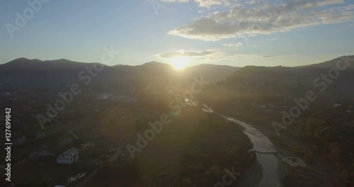 Aerial camera flies backwards over the village of Mykulychyn in the Carpathian region of Western Ukraine. The Prutets-Chemehivskyi river is below and the sun is rising behind distant mountains photo