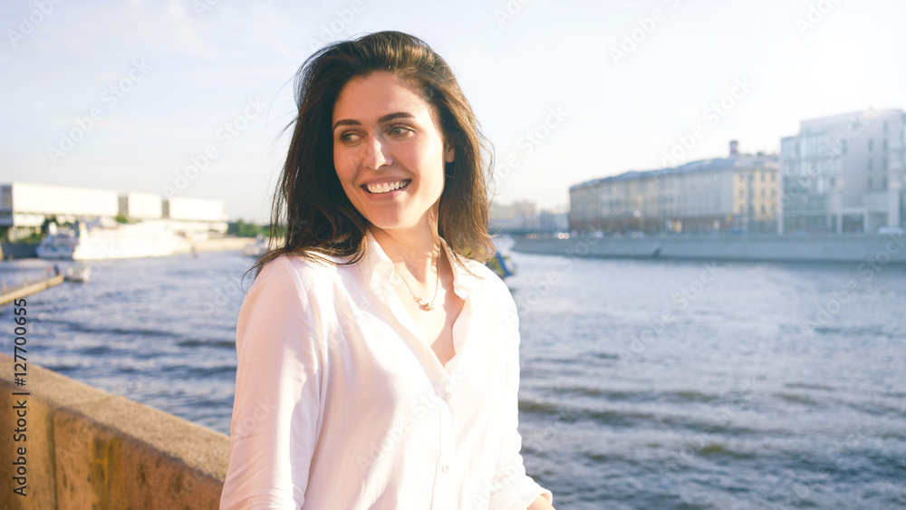 Young charming hipster girl with long brown hair is walking in the park on a river background. A beautiful brunette caucasian female wearing white shirt is having fun in a city park. Young charming hipster girl with long brown hair is walking in the park on a river background. A beautiful brunette caucasian female wearing white shirt is having fun in a city park.