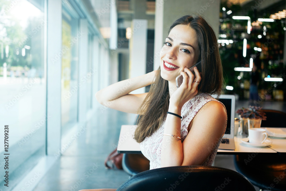 A charming female freelancer is talking on a phone while sitting in a cafe. Young attractive entrepreneur female is making a business call during the coffee break.