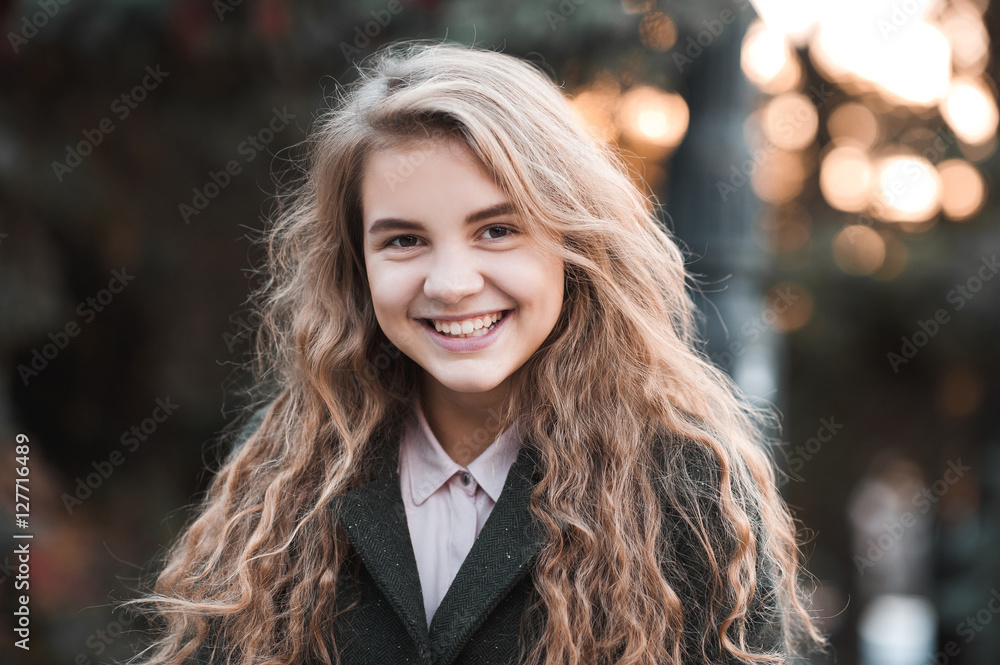 Obraz premium Smiling blonde teen girl 14-16 year old with curly hair wearing winter jacket over sunset at background. Looking at camera.