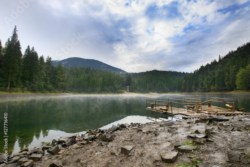 Mountain lake Synevir landscape with a mist low on water, two wooden floats at stony shore and coniferous forest around photo