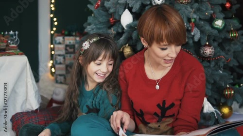 Mother with her daughter reading the book near dressed up christmas tree