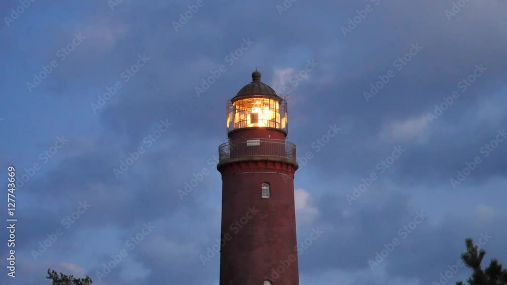 Shinning old lighthouse above pine forest after sunset. Tower illuminated with strong warning light. Lighthouse built from red bricks, gallery with iron handrail around glass cover of spotlight. Shinning old lighthouse above pine forest after sunset. Tower illuminated with strong warning light. Lighthouse built from red bricks, gallery with iron handrail around glass cover of spotlight.