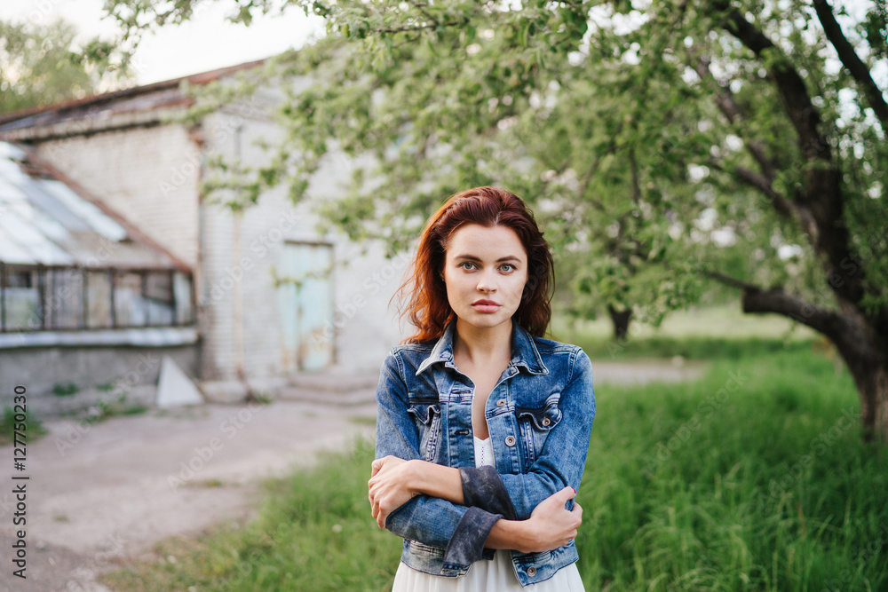 Obraz premium Young pretty serious caucasian woman in jeans jacket and white dress in a garden near the greenhouse. Youth, freshness, beauty concept.
