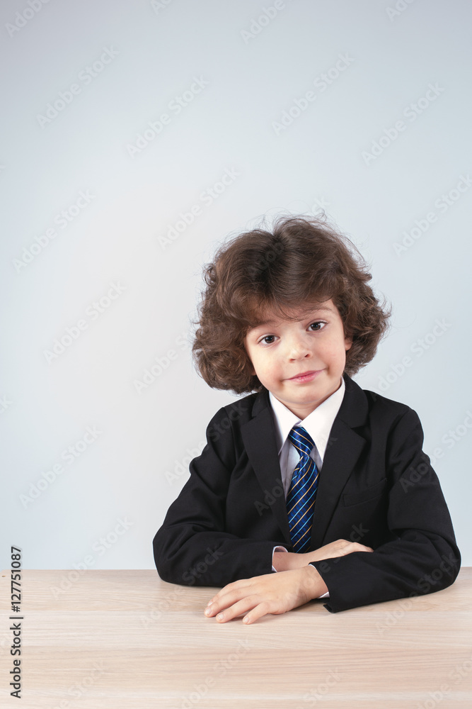 Cute little businessman sitting at the table and looking into the camera. Gray background. Cute little businessman sitting at the table and looking into the camera. Gray background.