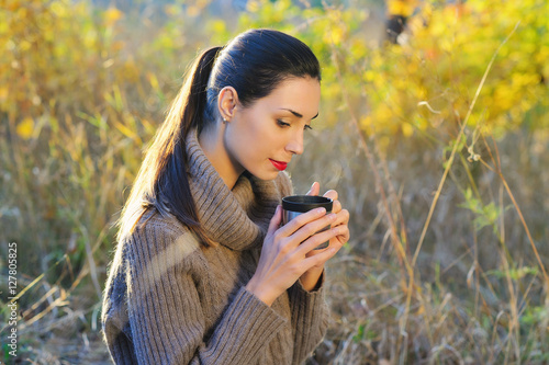 Beautiful young woman in autumn forest getting warm with cup of hot tea from thermos jug