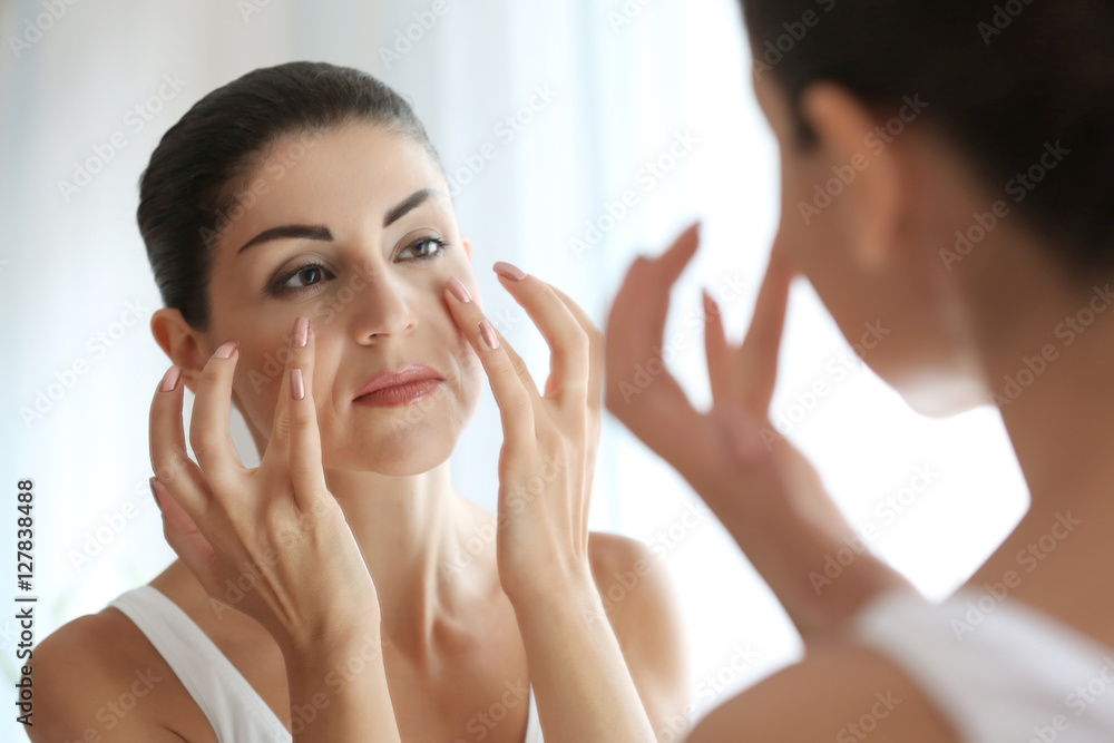 Adult woman checking her face in mirror on light background Adult woman checking her face in mirror on light background