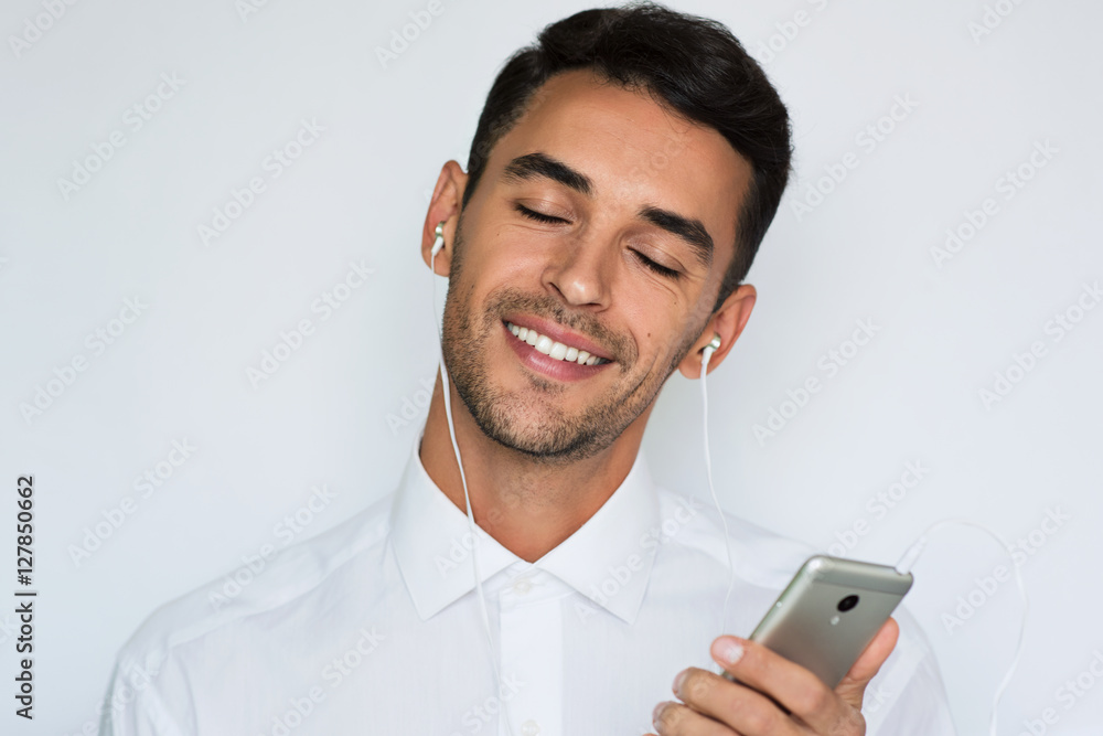 Beautiful portrait of a handsome attractive young smile man in white shirt with earphones holding mobile phone isolated on the light gray background. Happy businessman with smart phone listen music. Beautiful portrait of a handsome attractive young smile man in white shirt with earphones holding mobile phone isolated on the light gray background. Happy businessman with smart phone listen music.