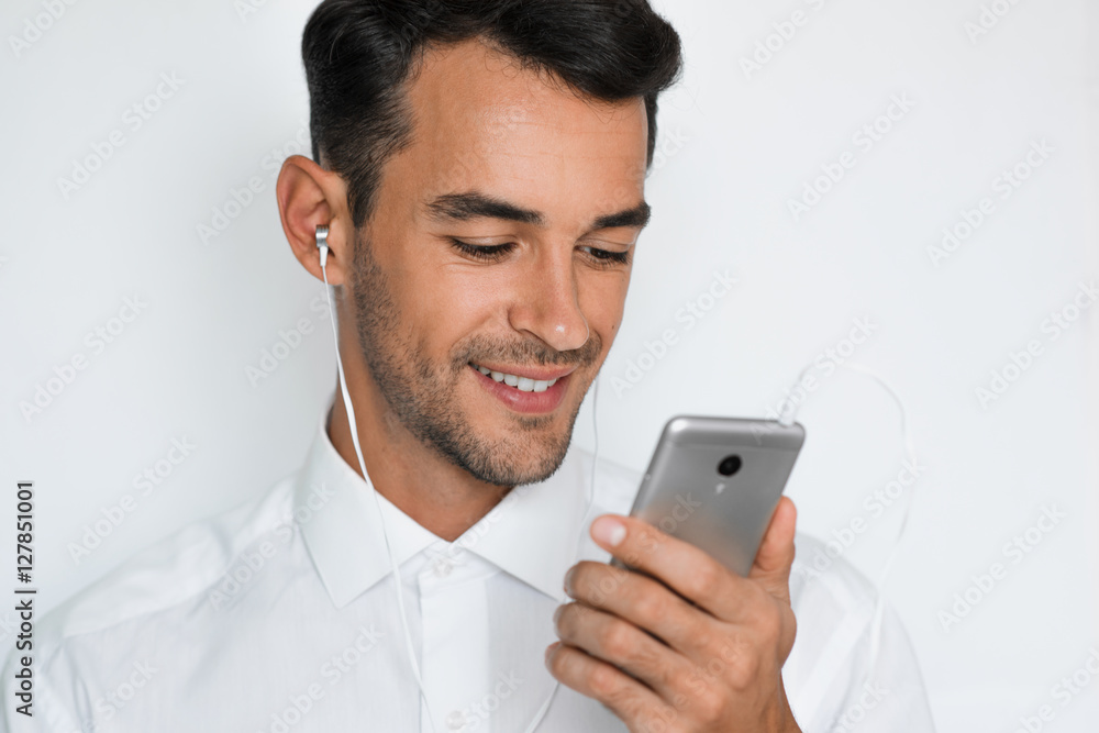 Portrait of a handsome attractive young man in white shirt with earphones holding mobile phone isolated on the light gray background. Happy businessman with smart phone listen trendy concept. Portrait of a handsome attractive young man in white shirt with earphones holding mobile phone isolated on the light gray background. Happy businessman with smart phone listen trendy concept.