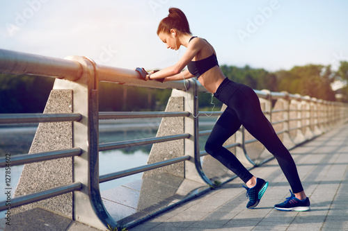 Attractive and strong woman stretching before fitness and listening to music with headphones on the lake in the summer. Sports concept. Healthy lifestyle