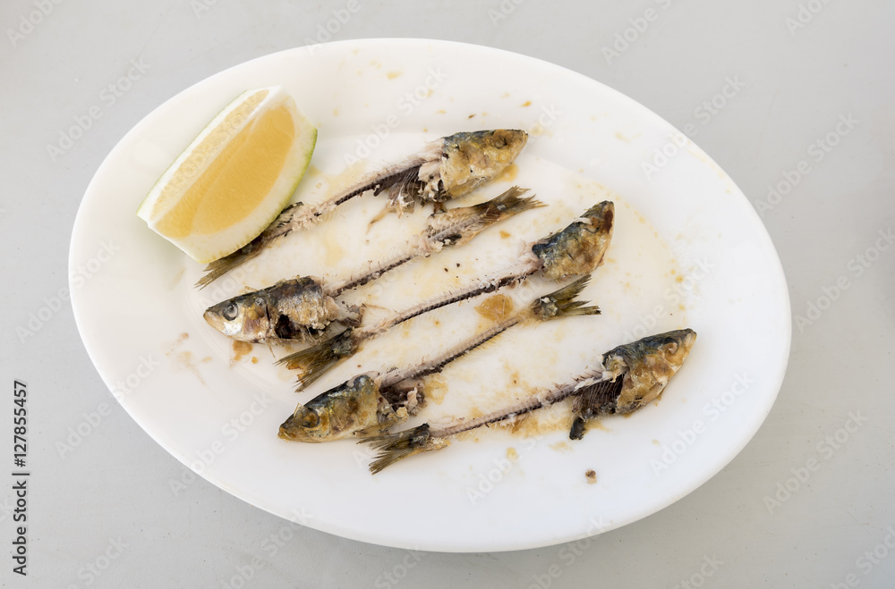 Bones of five eaten Sardines, on a white plate. with the head and tails intact, and a wedge of Lemon on the side Bones of five eaten Sardines, on a white plate. with the head and tails intact, and a wedge of Lemon on the side