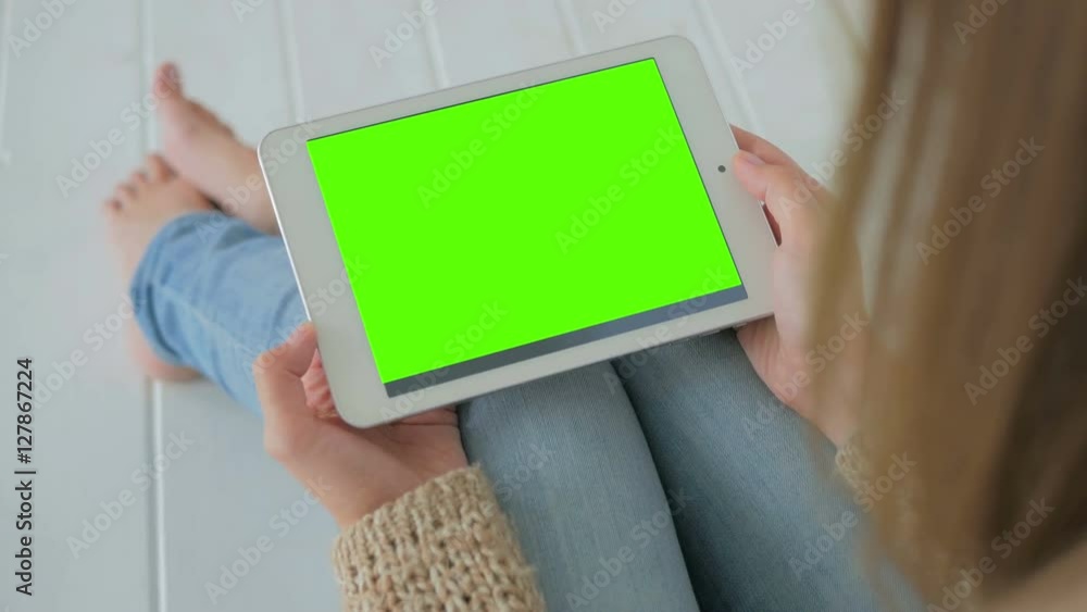 Woman sitting on the white wooden floor and looking at horizontal tablet computer with green screen. Close up shot of woman's hands with pad Woman sitting on the white wooden floor and looking at horizontal tablet computer with green screen. Close up shot of woman's hands with pad