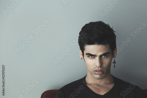 Portrait of attractive modern young white brazilian guy with black hair and cross-earring looking sullenly and saucily, gray wall behind photo